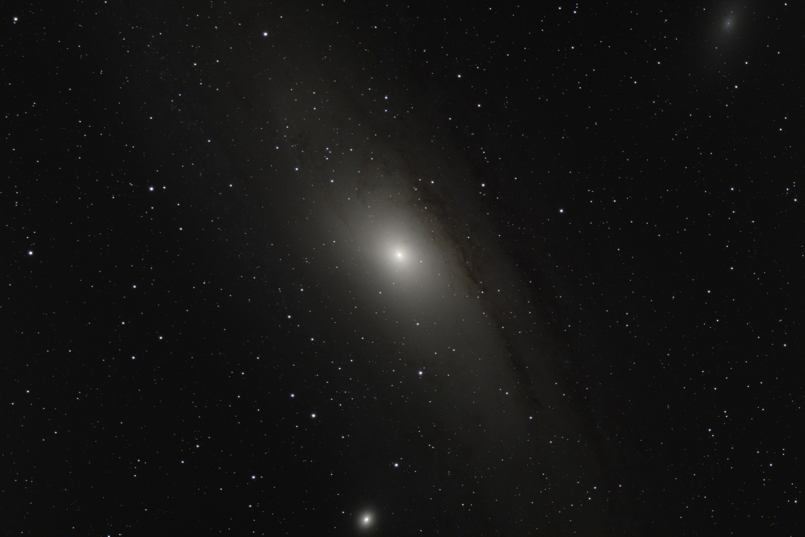 The core of the Andromeda galaxy filling the frame at a tilt: a bright nucleus, a broad glowing bulge, and dark dust lanes combed through the far side of the disk, with the small companion galaxy M32 at the bottom edge and M110 in the upper right corner.