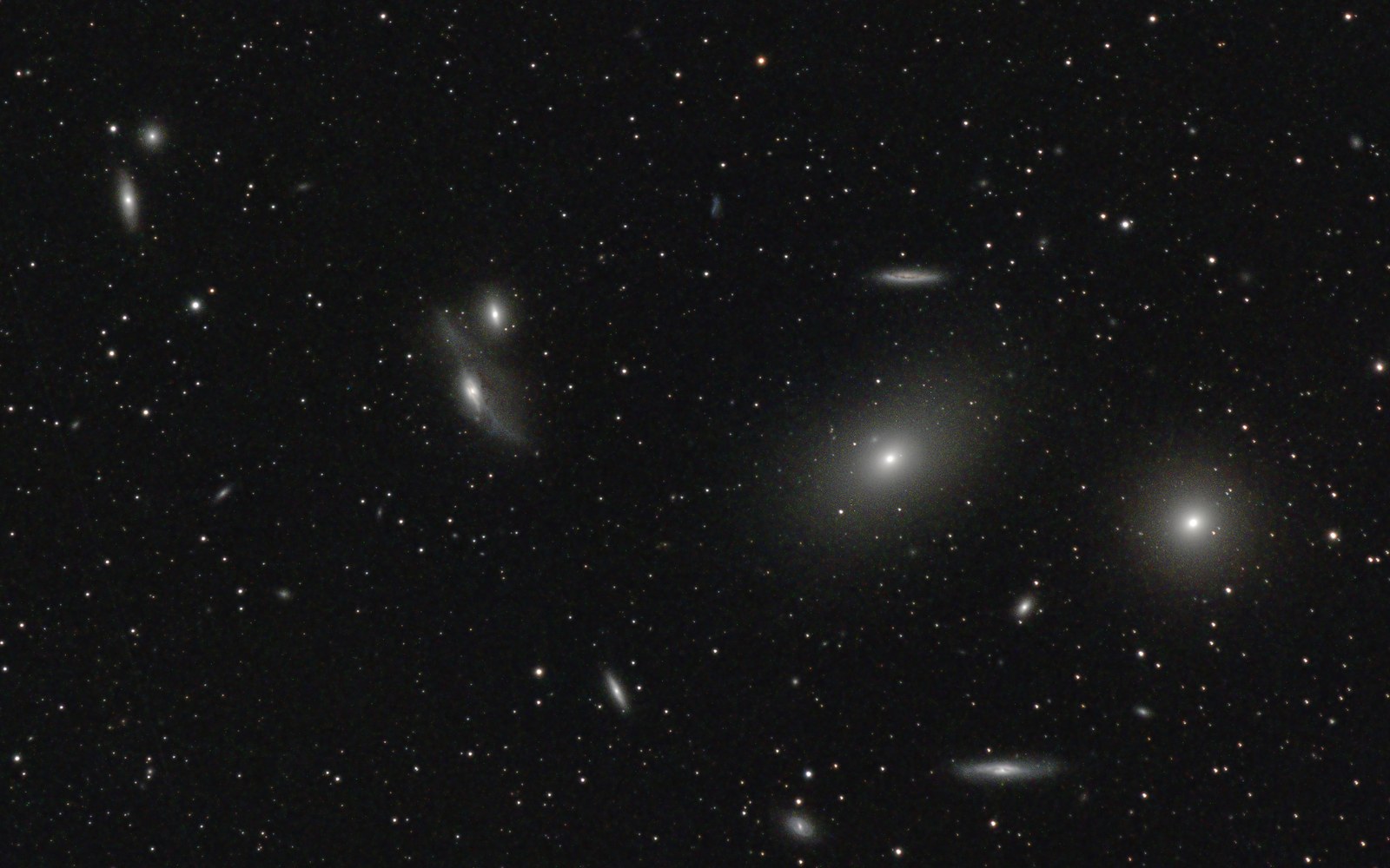 A dense field of small galaxies: two large soft elliptical glows on the right, a close pair of interacting galaxies left of center with a faint bridge between them, several edge-on spirals like thin streaks, and dozens of fainter smudges among the stars.